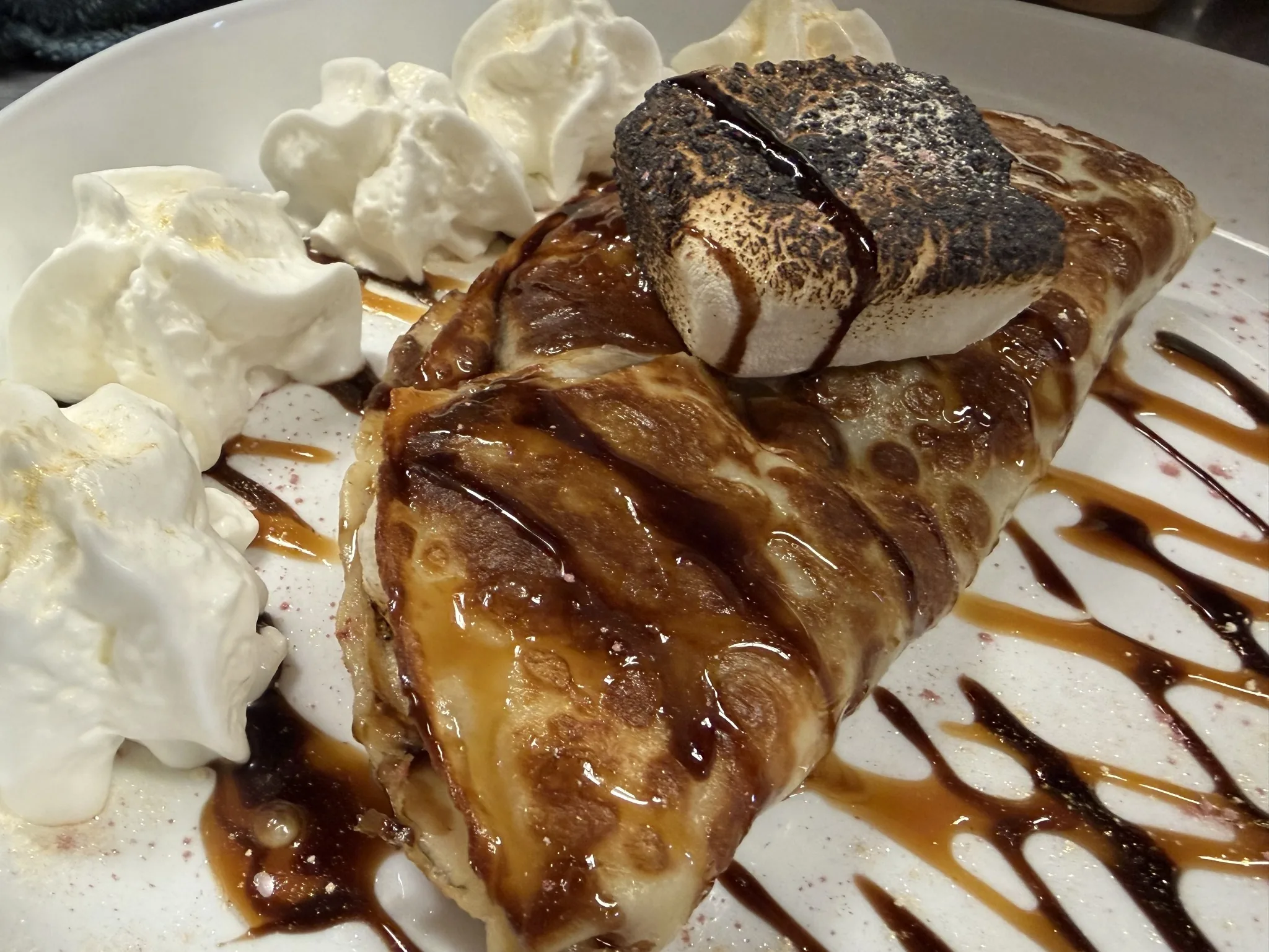 Dessert crepe filled with Nutella and topped with a toasted marshmallow and served with whipped cream, chocolate drizzle, and caramel on a white plate at Kat’s Café & Restaurant.