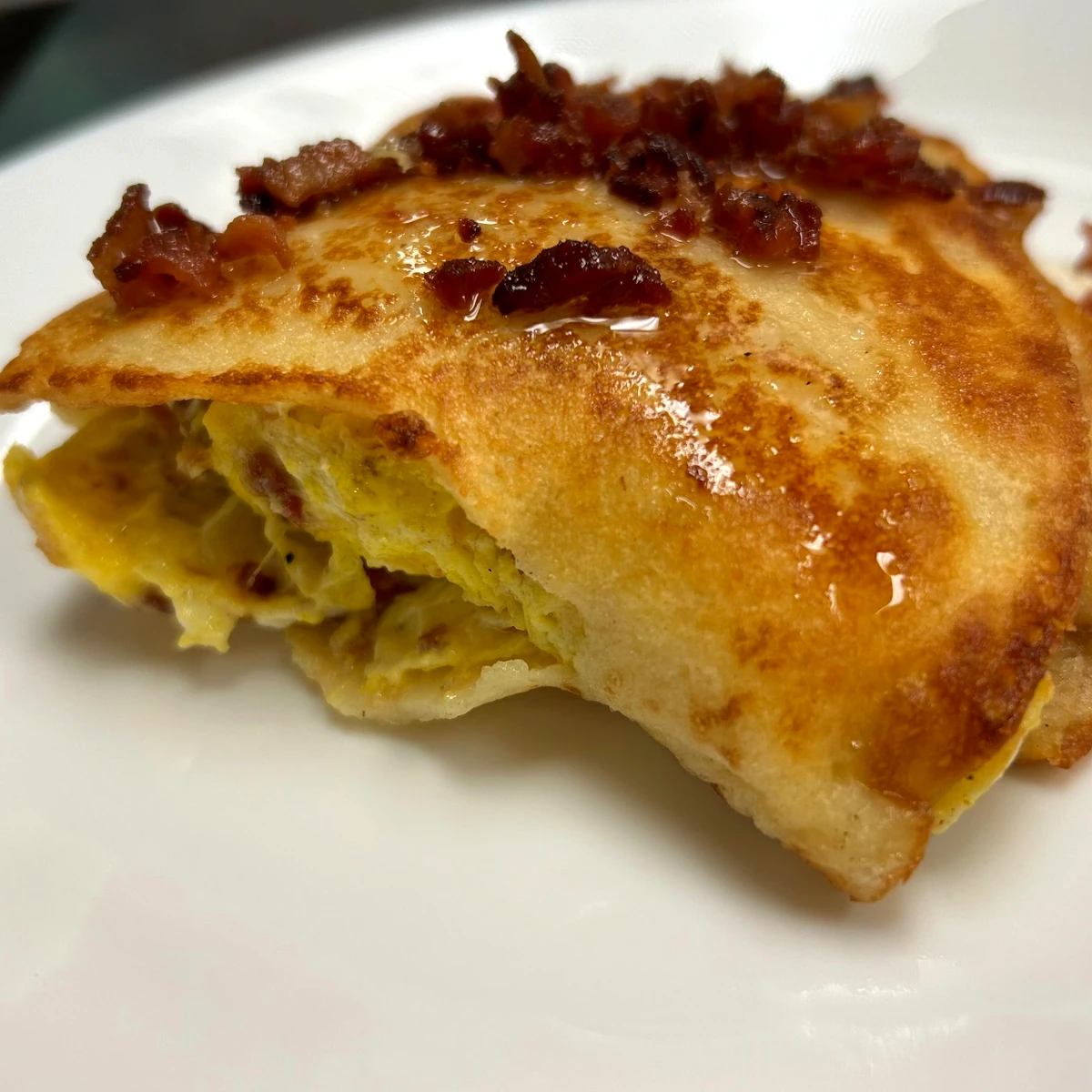 Golden-brown bacon, egg, and cheese blintz topped with crispy bacon pieces on a white plate.