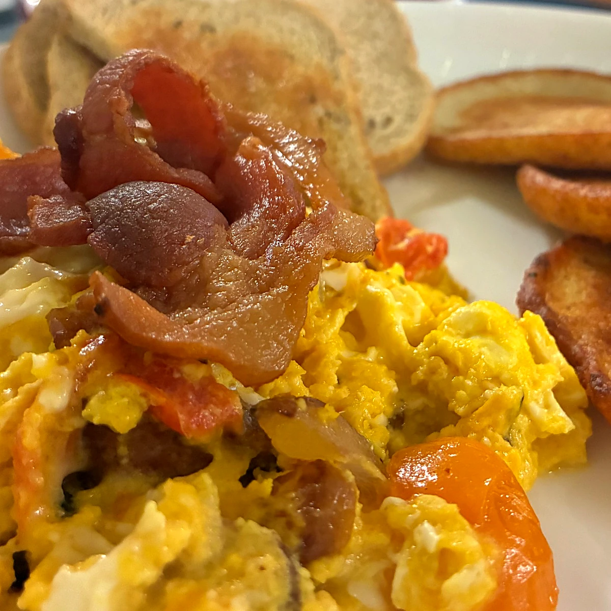 Scrambled eggs having cherry tomatoes and topped with crispy bacon, served with golden blinis and toast on a white plate.