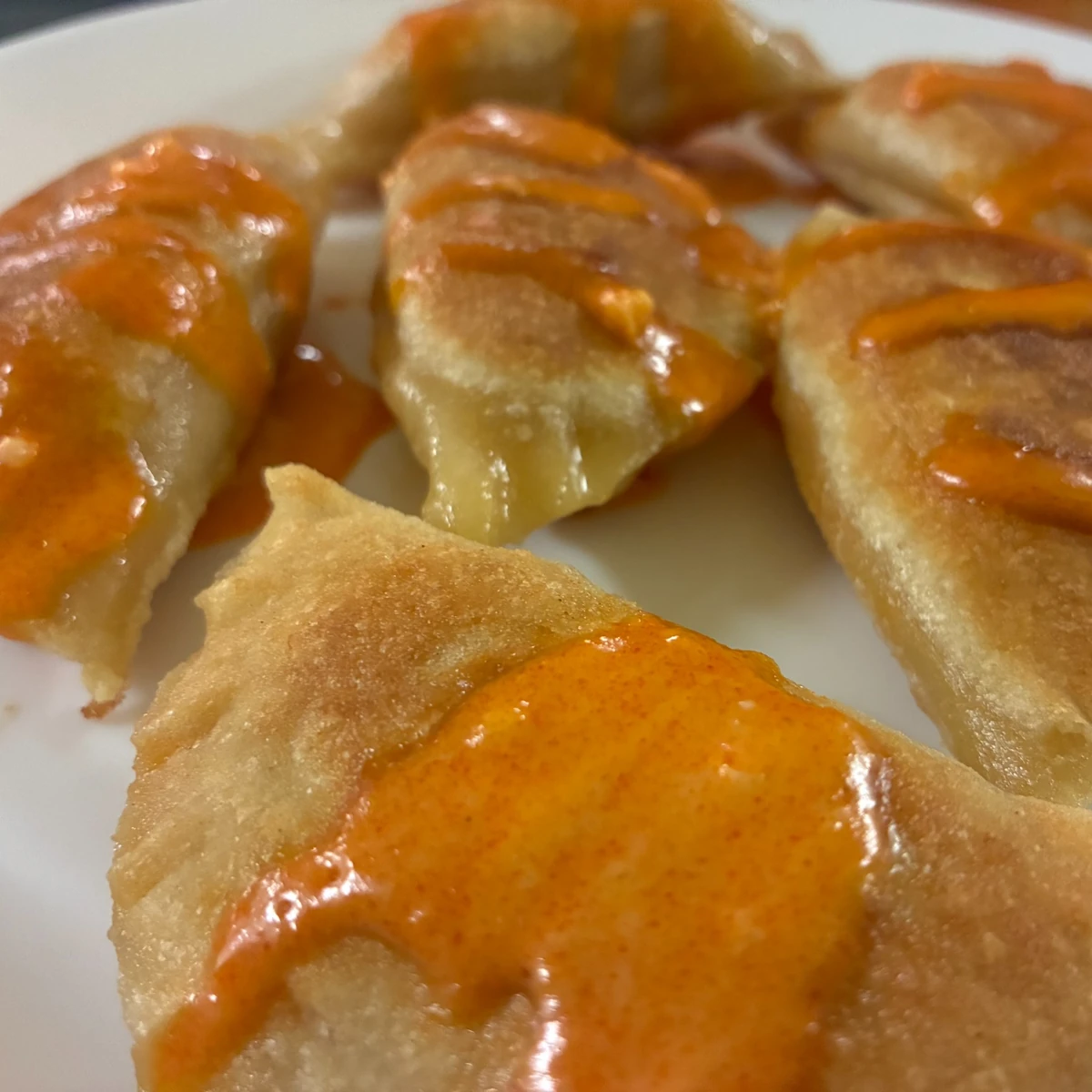 Pan-seared pierogi drizzled with orange buffalo-style sauce on a white plate.
