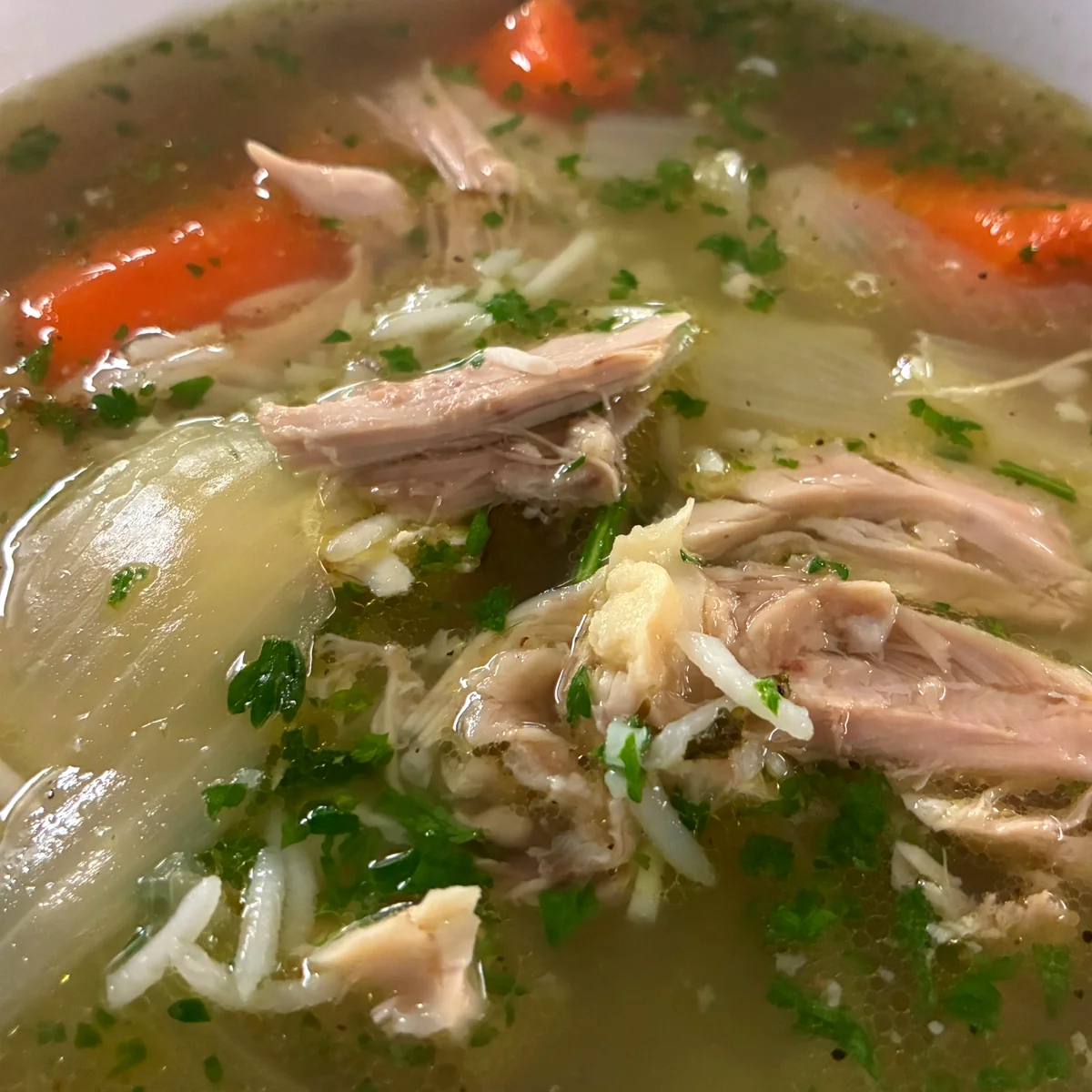 Close-up of clear chicken soup (rosół) with tender chicken, carrots, rice, onion, and fresh herbs in a white bowl.