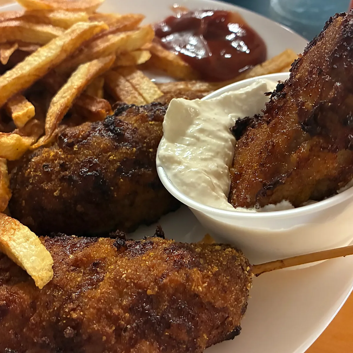 Breaded city chicken skewers with fries and a cup of creamy dipping sauce on a white plate.