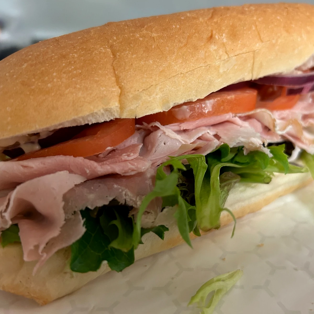 Off the bone country ham sub on soft hoagie bread with lettuce, tomato slices, mayo, and red onion on takeout wrapping paper.