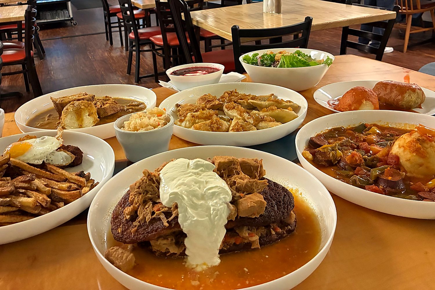 Assorted dinner plates and comfort-food spread on a wooden table with pierogi, borscht, salad, fries, and hearty mains in Kat’s dining room.