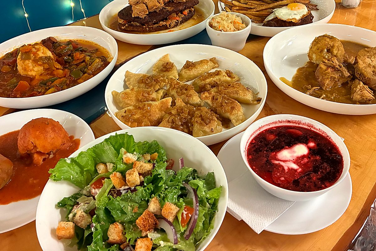 Family-style comfort-food spread on a wooden table with pierogi, borscht, salad, cutlet, fries, and hearty mains, with soft string-light bokeh in the background at Kat’s Café & Restaurant.