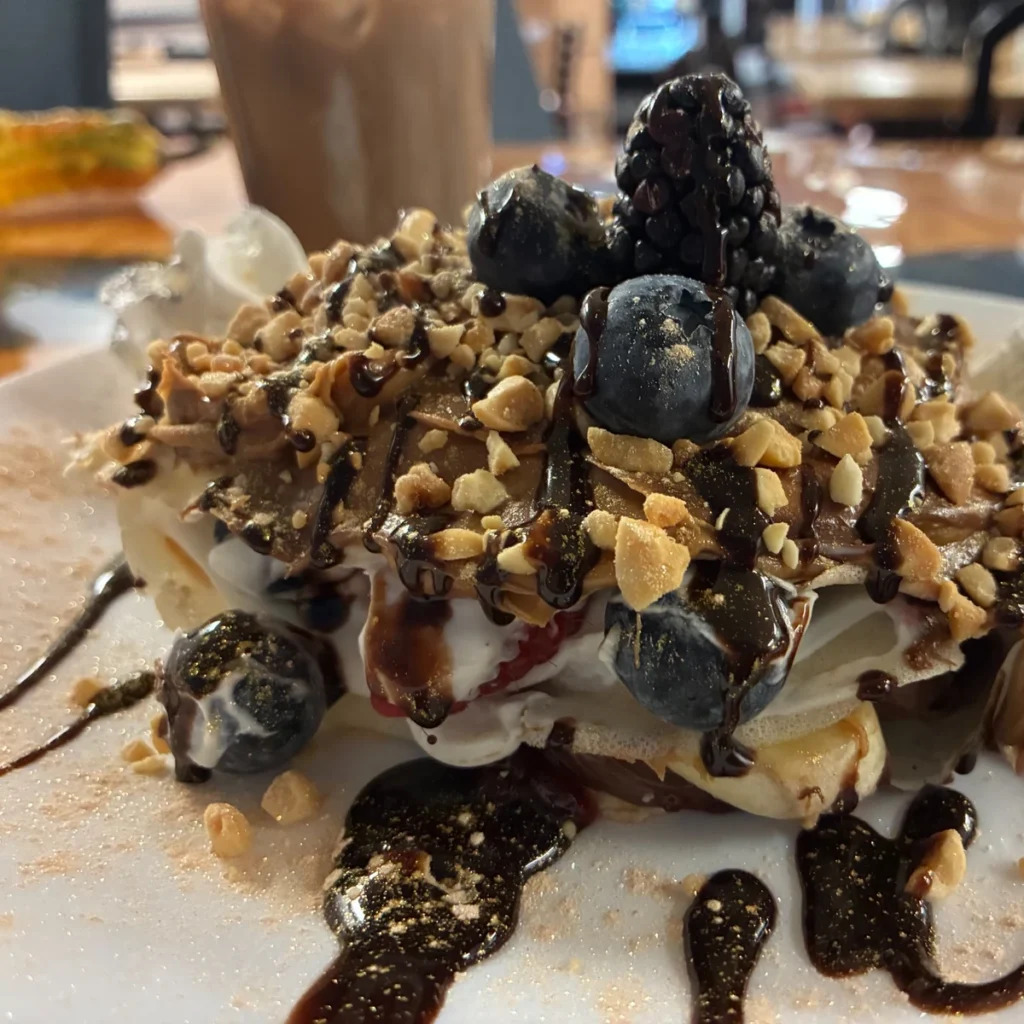 Loaded dessert crepe blintz topped with whipped cream, blueberries, a blackberry, crushed nuts, and chocolate drizzle on a plate, with an iced coffee blurred in the background.