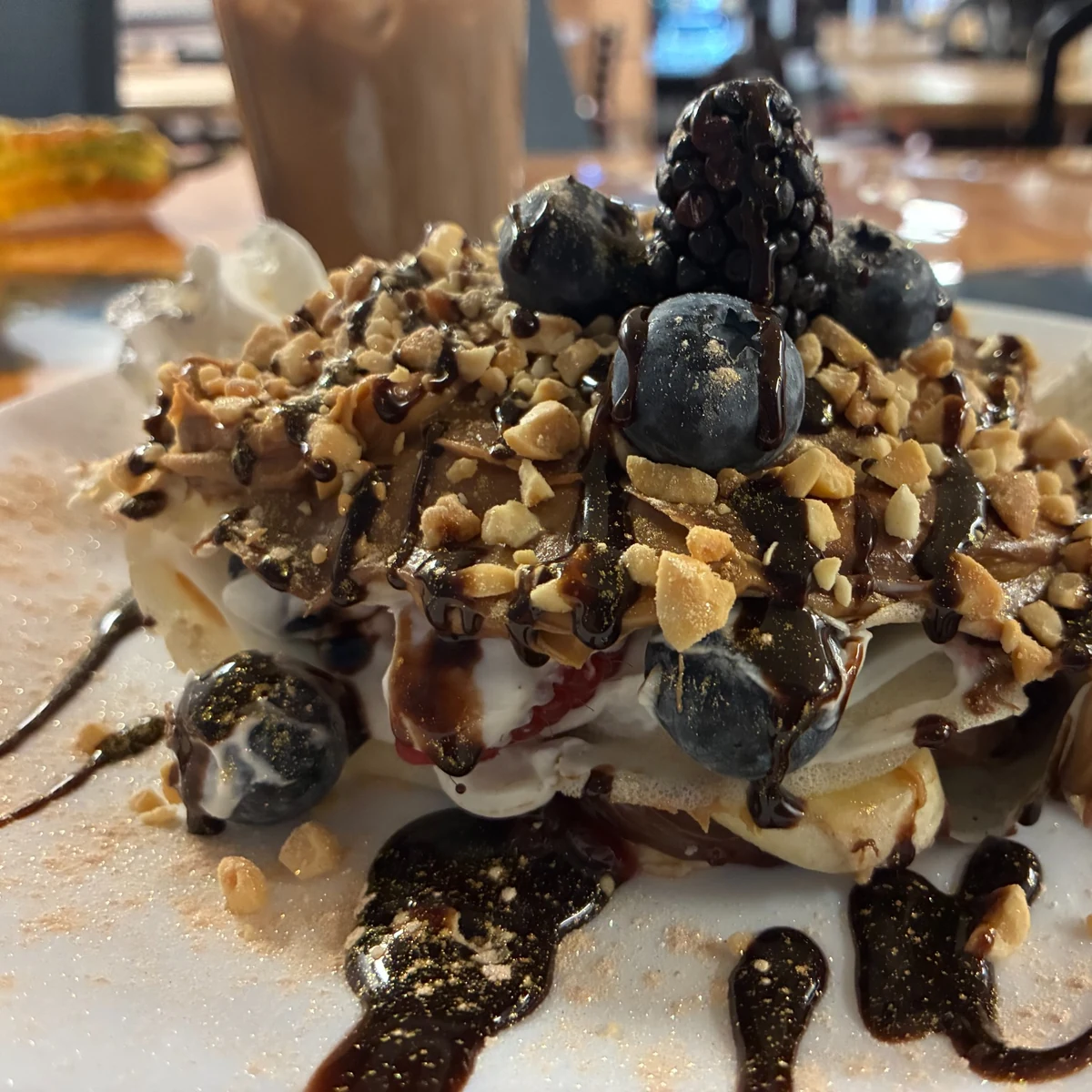 Loaded dessert crepe blintz topped with whipped cream, blueberries, a blackberry, crushed nuts, and chocolate drizzle on a plate, with an iced coffee blurred in the background.