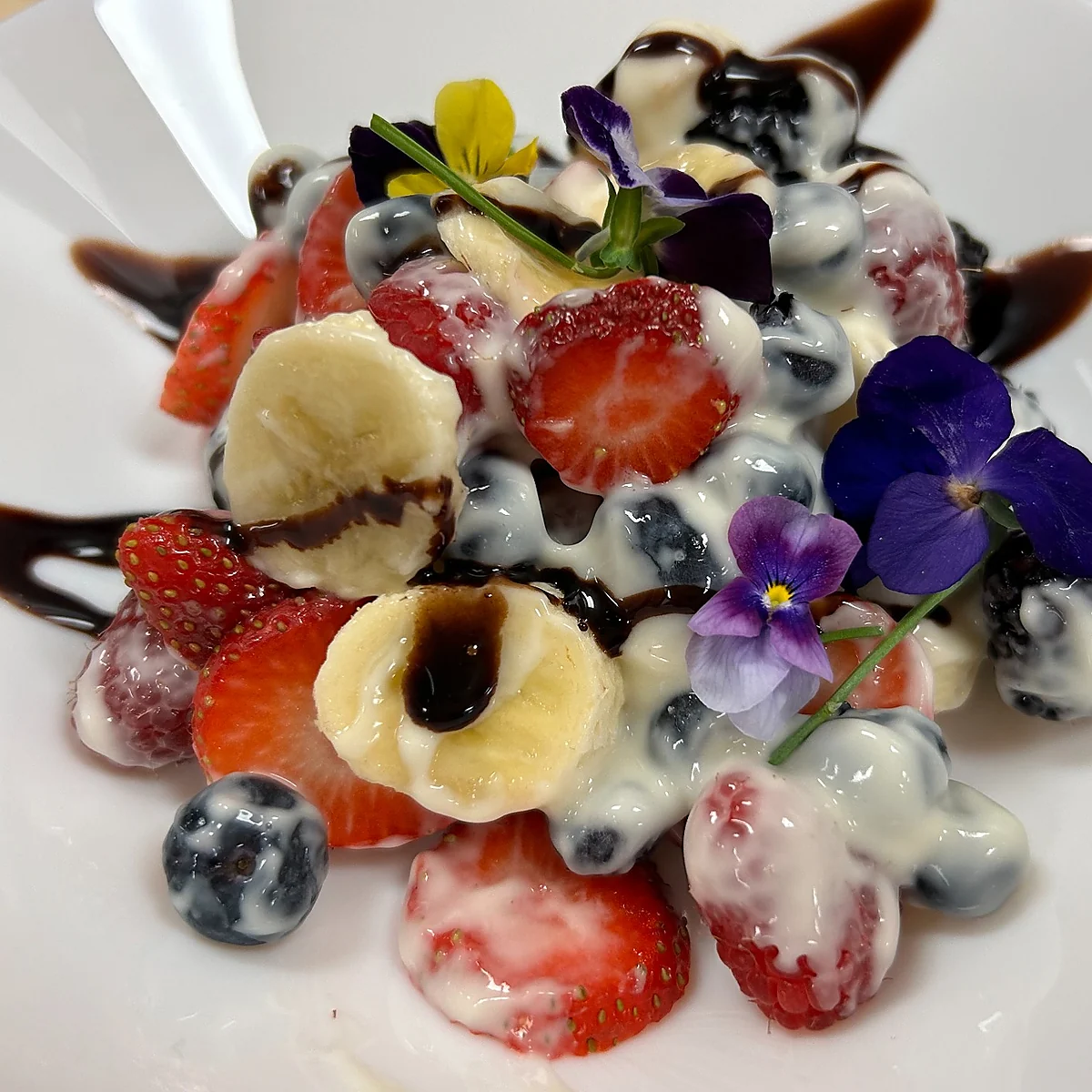 Fruit salad with strawberries, blueberries, raspberries, banana slices, sweet cream, chocolate drizzle, and edible flowers on a white plate.