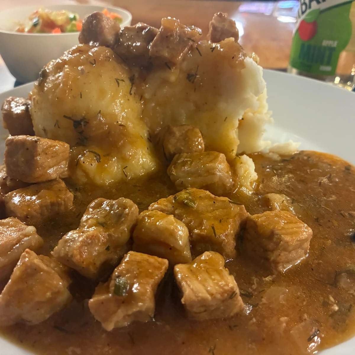 Tender goulash with gravy served alongside mashed potatoes on a plate with a veggie salad and apple mint juice in the background.