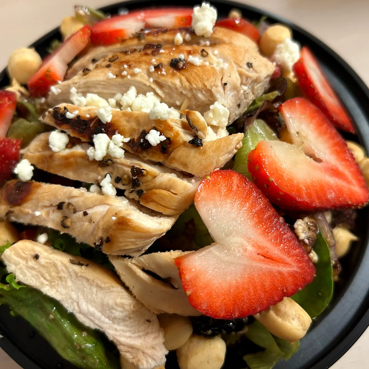 Grilled chicken salad with sliced strawberries, berries, nuts, and crumbled cheese in a black bowl.