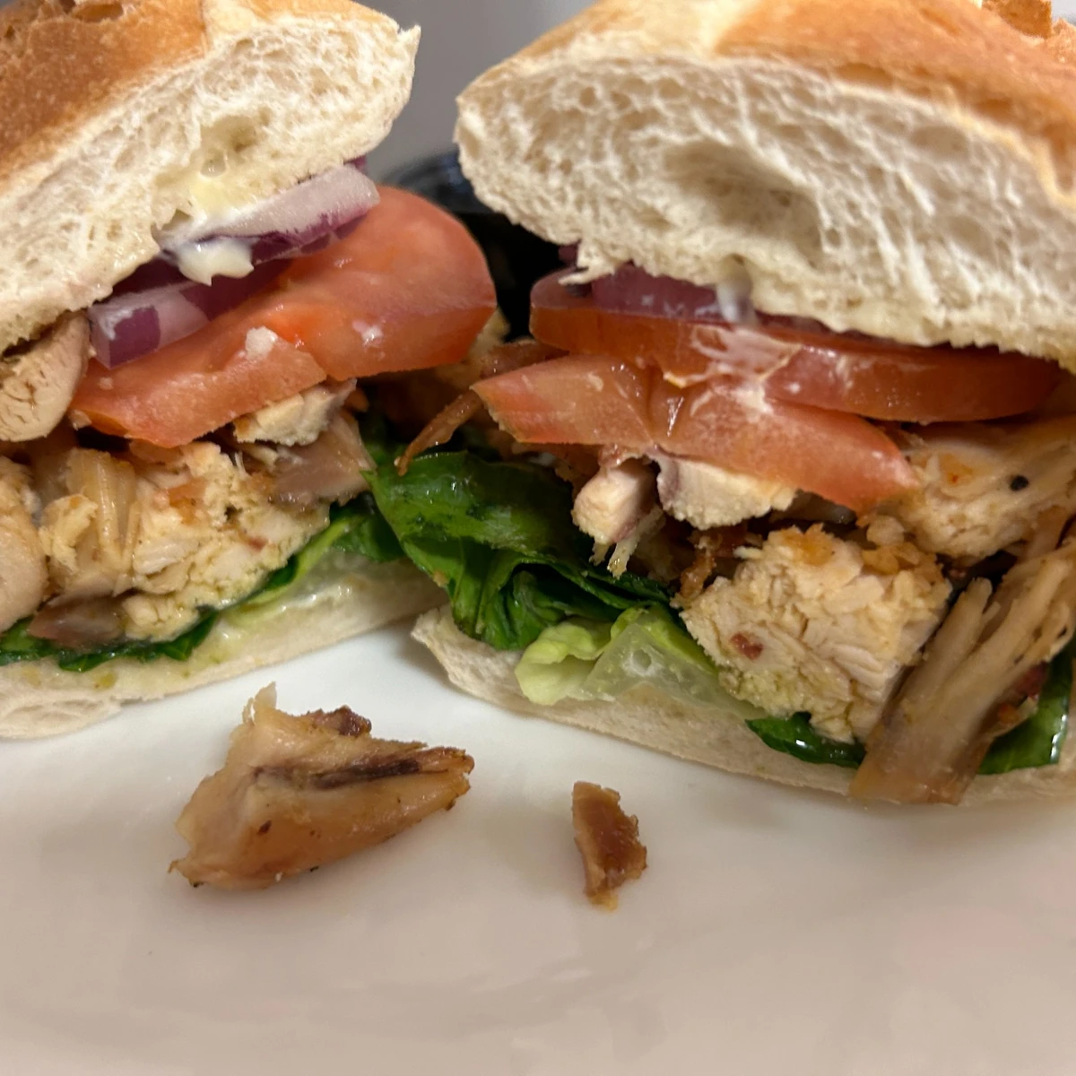 Grilled chicken sub cut in half on a white plate, layered with tomatoes, red onion, mayo, and leafy greens.
