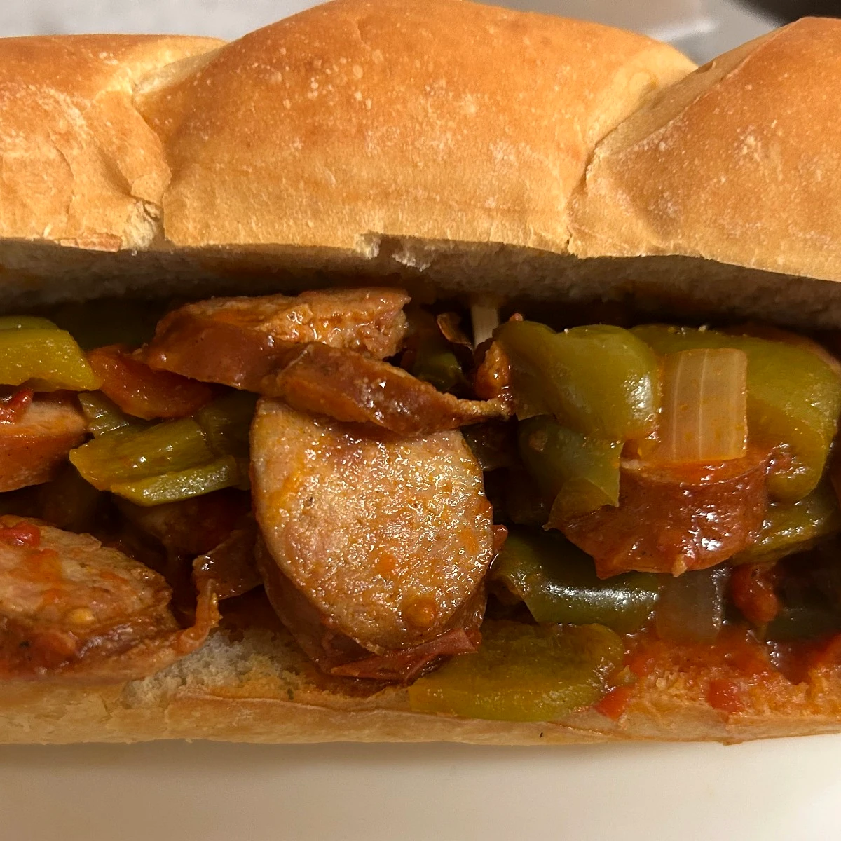Kielbasa hoagie loaded with sautéed peppers, stewed tomatoes, and onions on a white plate.