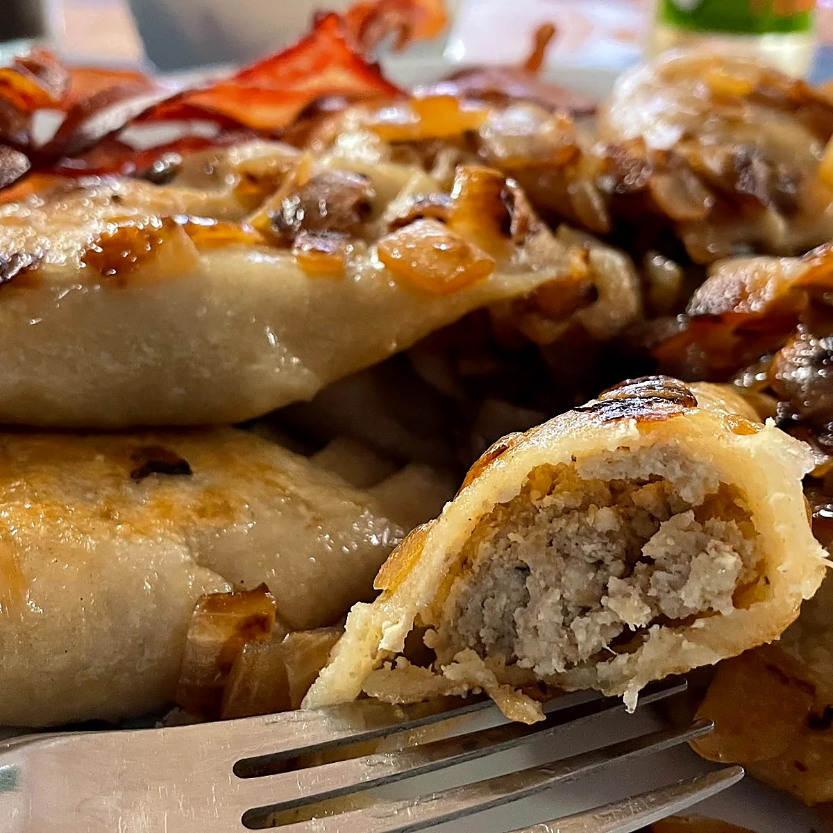 Meat-filled pierogi topped with fried onions and bacon, with one pierogi opened to show the filling.