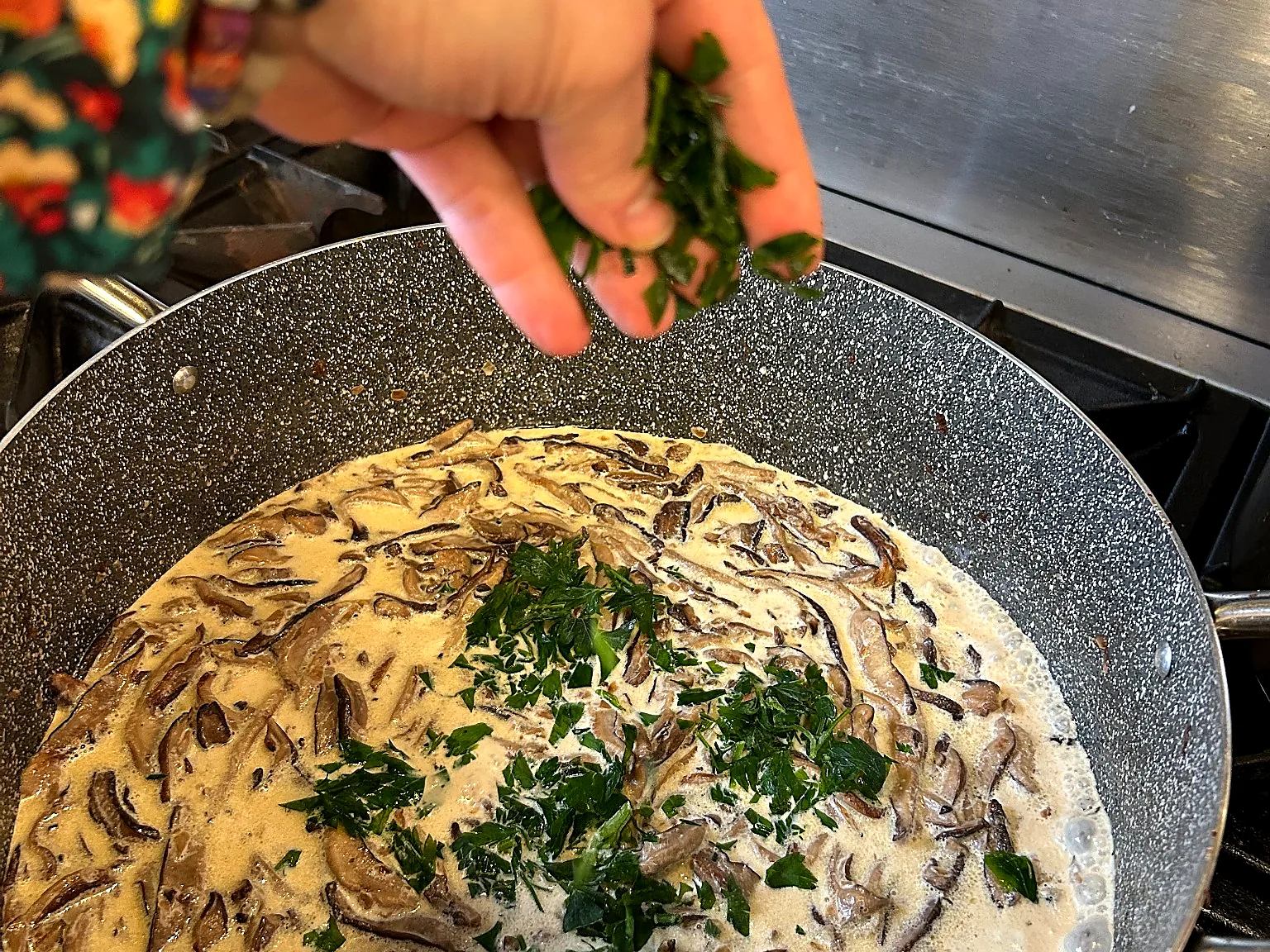 Hand sprinkling fresh herbs into creamy mushroom sauce in a pan.