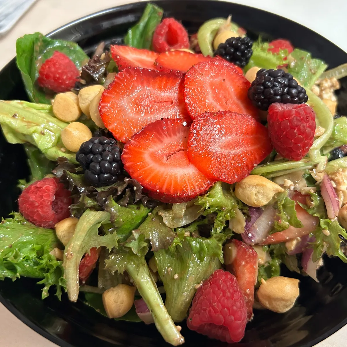 Mixed greens salad topped with strawberries, raspberries, blackberries, puffy croutons, and nuts in a black bowl.