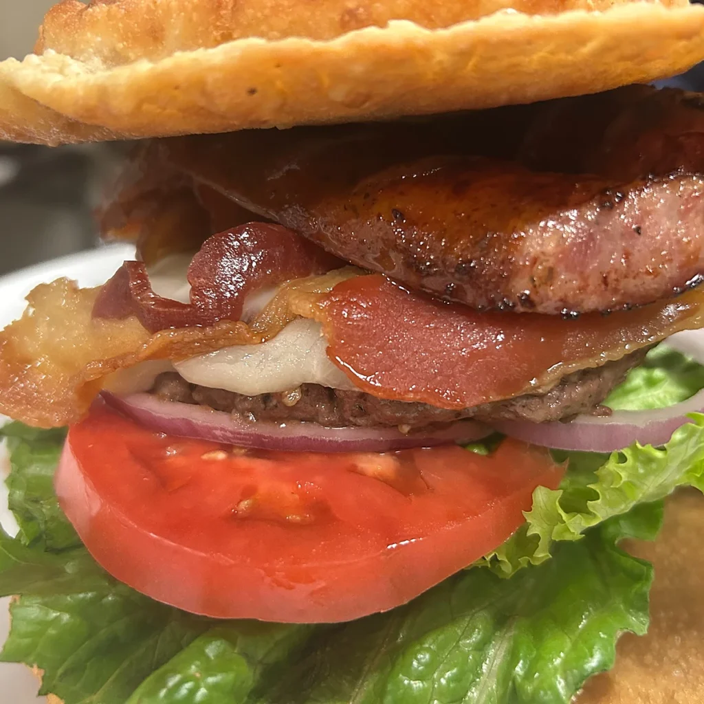 Close-up of a pierogi-bun smash burger topped with a thick slice of kielbasa and crispy bacon, layered with tomato, red onion, and lettuce.