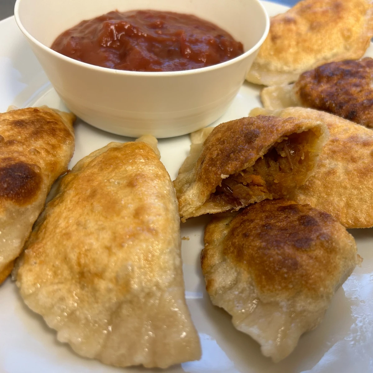 Pan-fried seasonal autumn harvest pierogi with a bite showing orange-brown filling and a bowl of red holiday dipping sauce nearby.