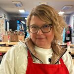 Chef Kat in a red apron seated at the custom wooden table in Kat's cozy dining room at her cafe-restaurant in Minersville, PA.