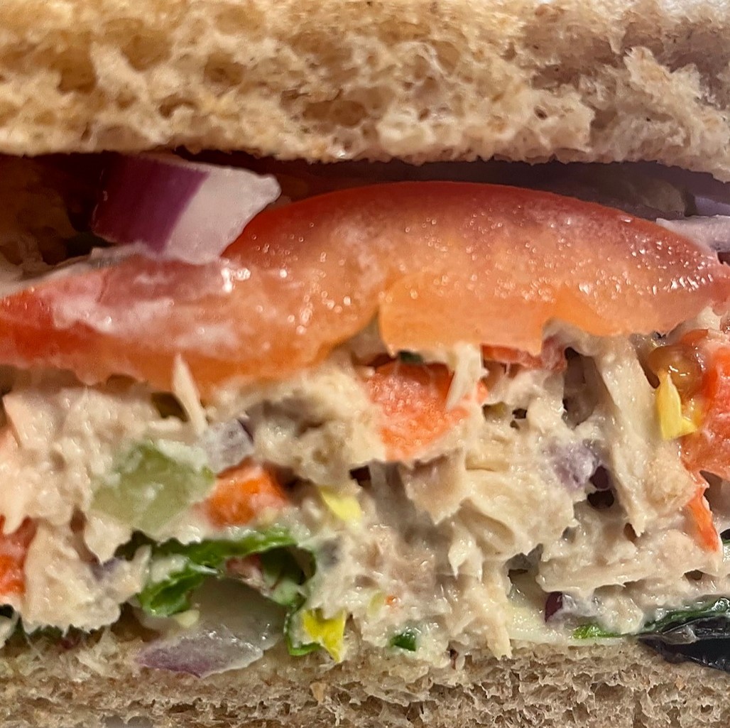 Close-up of a tuna salad sub with tiny veggie pieces, sliced tomato, red onion, and greens on fresh bread.