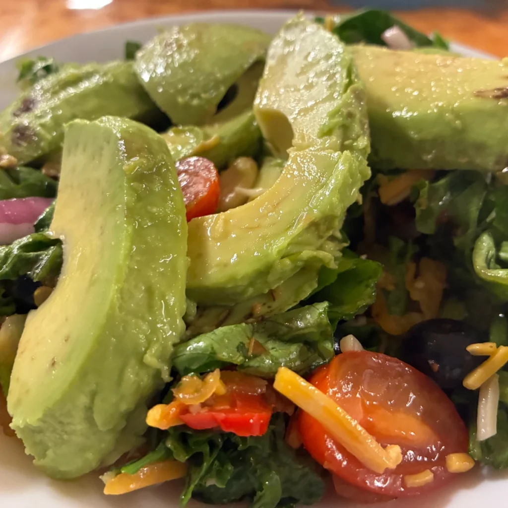Avocado slices over mixed greens with cherry tomatoes and shredded cheese on a white plate.