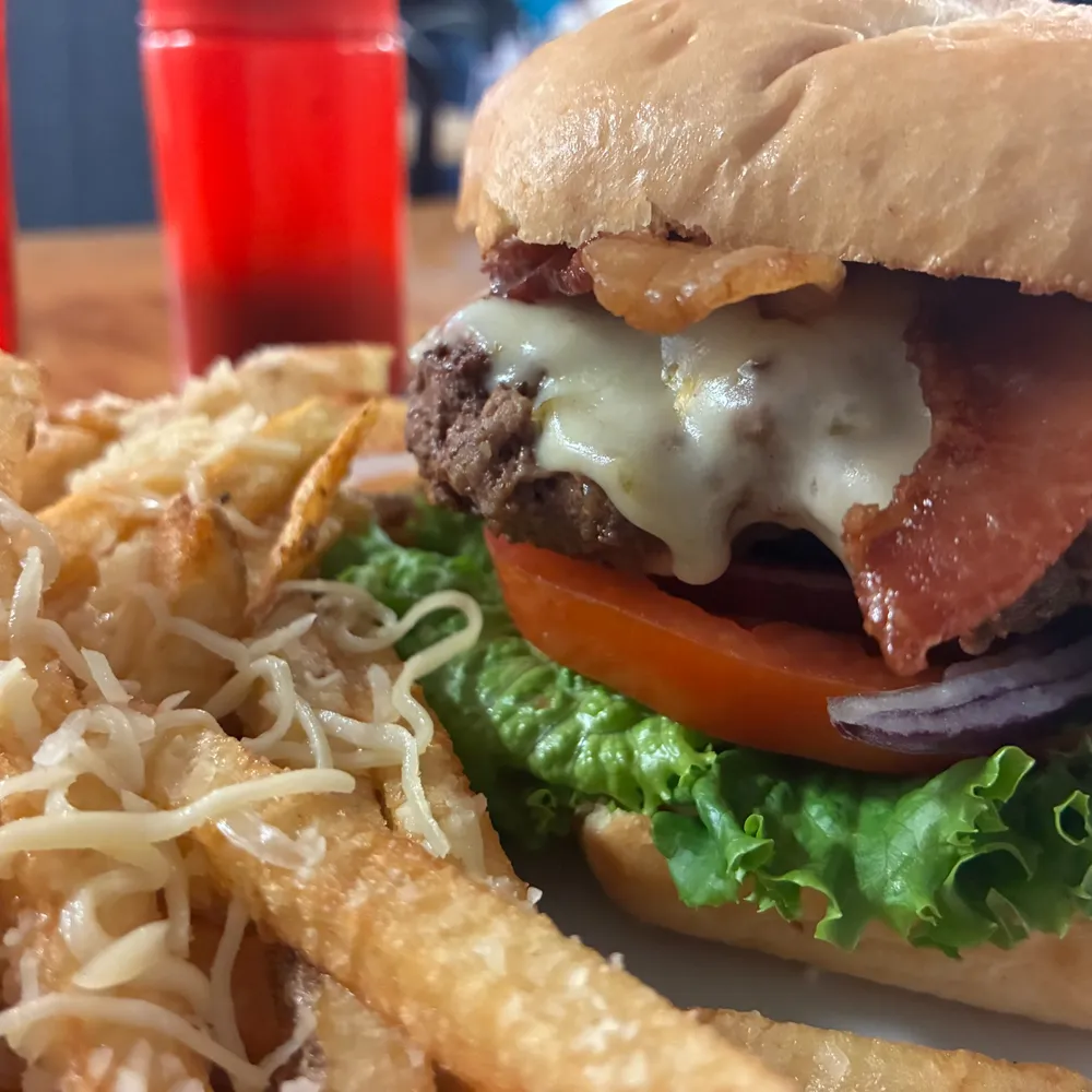 Bacon cheeseburger with lettuce, tomato and red onion, served with Parmesan fries and a ketchup bottle and glass of juice in the background.
