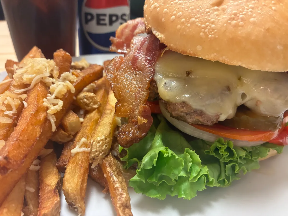 Close-up of a bacon cheese smash burger with lettuce, tomato and fries topped with Parmesan on a white plate.