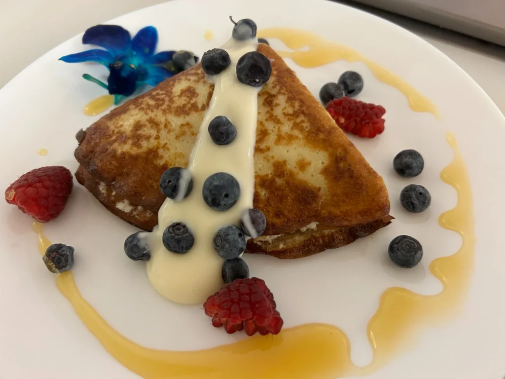 Sweet cheese blintz crepe topped with blueberries, raspberries and a creamy white sauce on a white plate garnished with caramel sauce, edible flowers, and berries.