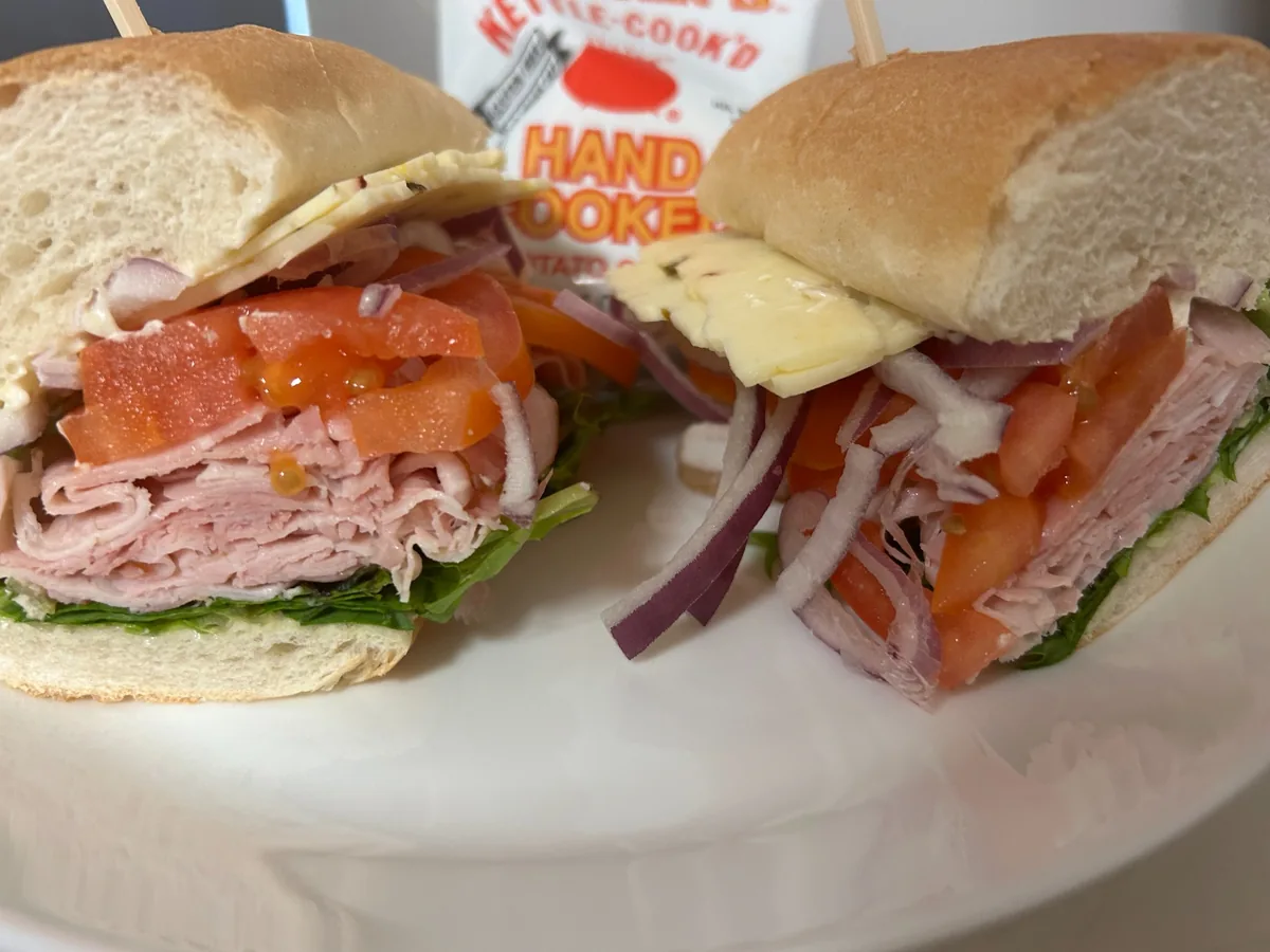 Country ham sub cut in half with sliced off-the-bone ham, tomato, red onion, lettuce, and pepper jack cheese on soft hoagie bread served on a white plate with a bag of chips in the background.