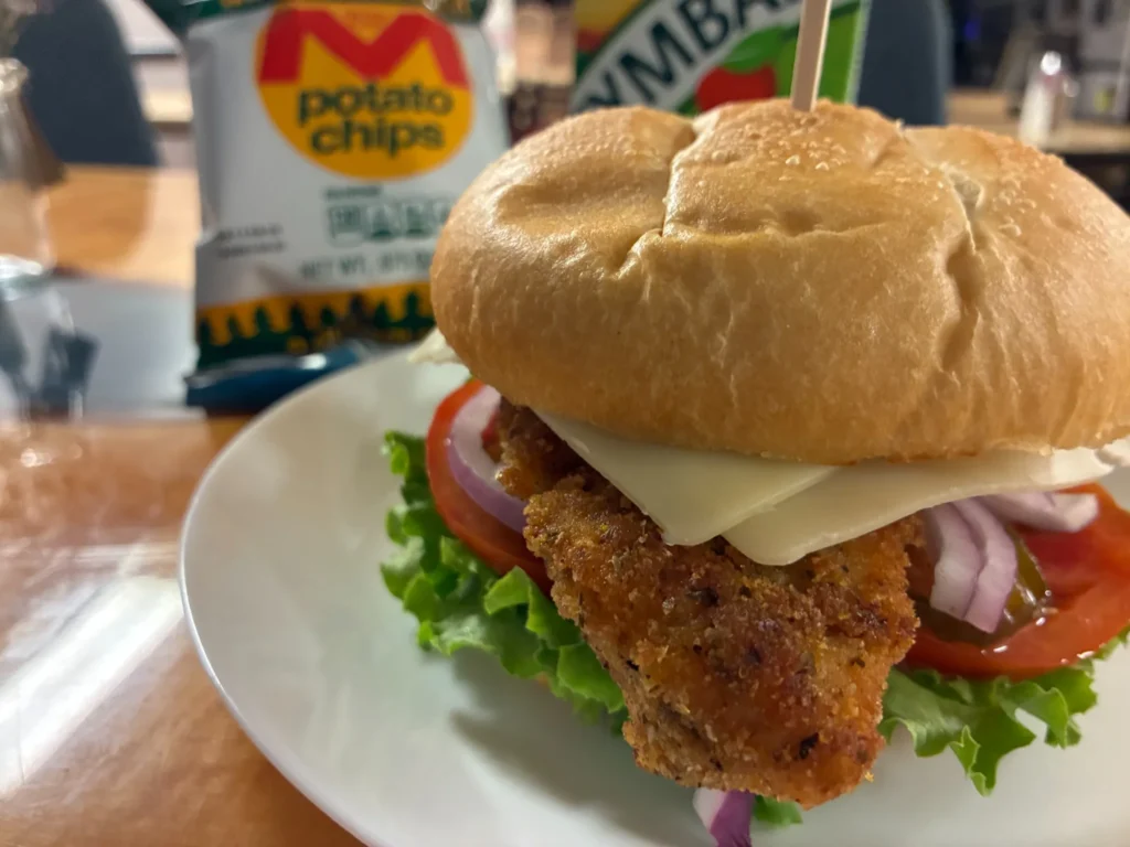Breaded chicken cutlet sandwich on a fresh roll with lettuce, tomato, red onion and yellow cheese with a bag of chips and fruit juice in the background.