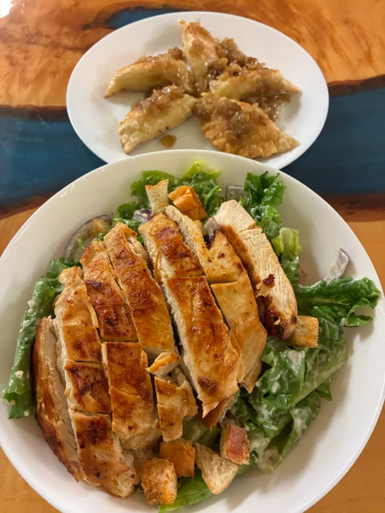 Chicken Parmesan Caesar salad topped with sliced grilled chicken and croutons, served with a plate of pierogi in the background.