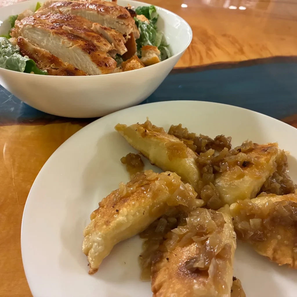 Plate of pan-fried pierogies topped with caramelized onions on a table, with a chicken Caesar salad in a bowl behind.