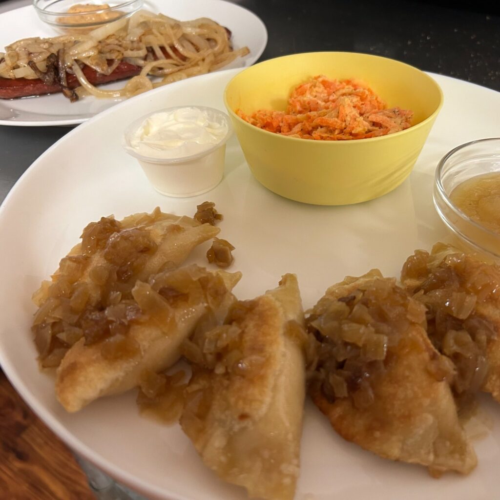 Pan-fried pierogies topped with onions served with sour cream, applesauce, and carrot salad on a white platter with fried kielbasa and onions on a plate behind it.