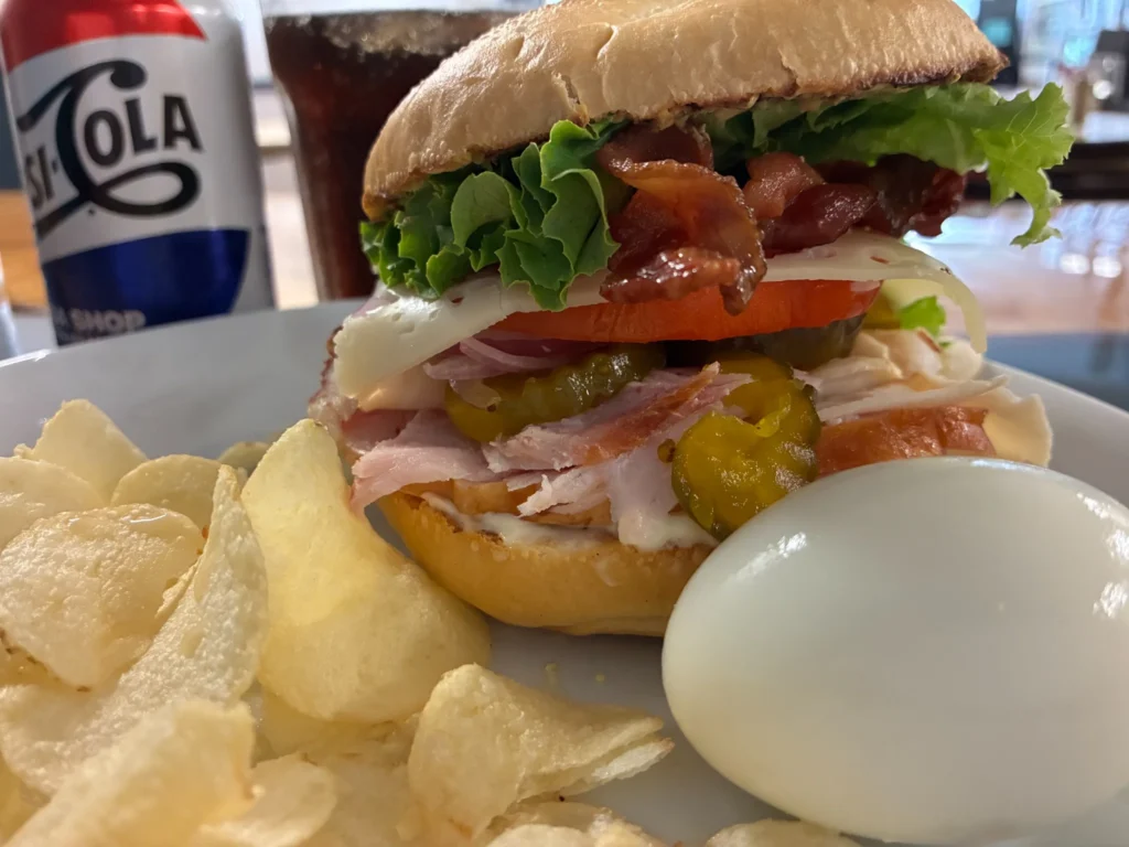 Country ham sandwich topped with bacon, cheese, pickles, lettuce and tomato, served with chips and a hard-boiled egg.