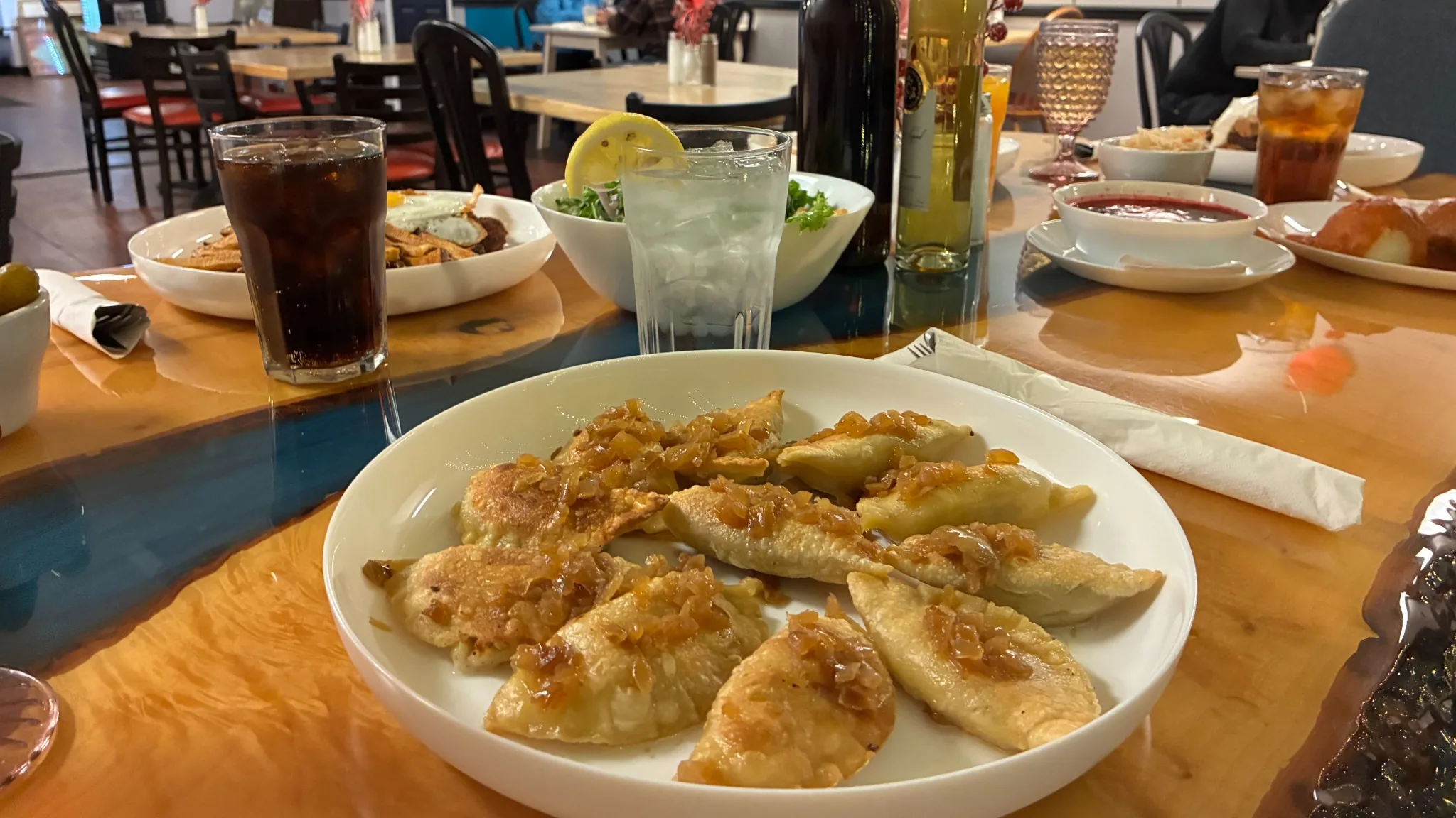 Pan-fried pierogies topped with caramelized onions served on a white platter on a table with drinks, other plates, and wine, with diners and Kat's dining area in the background.