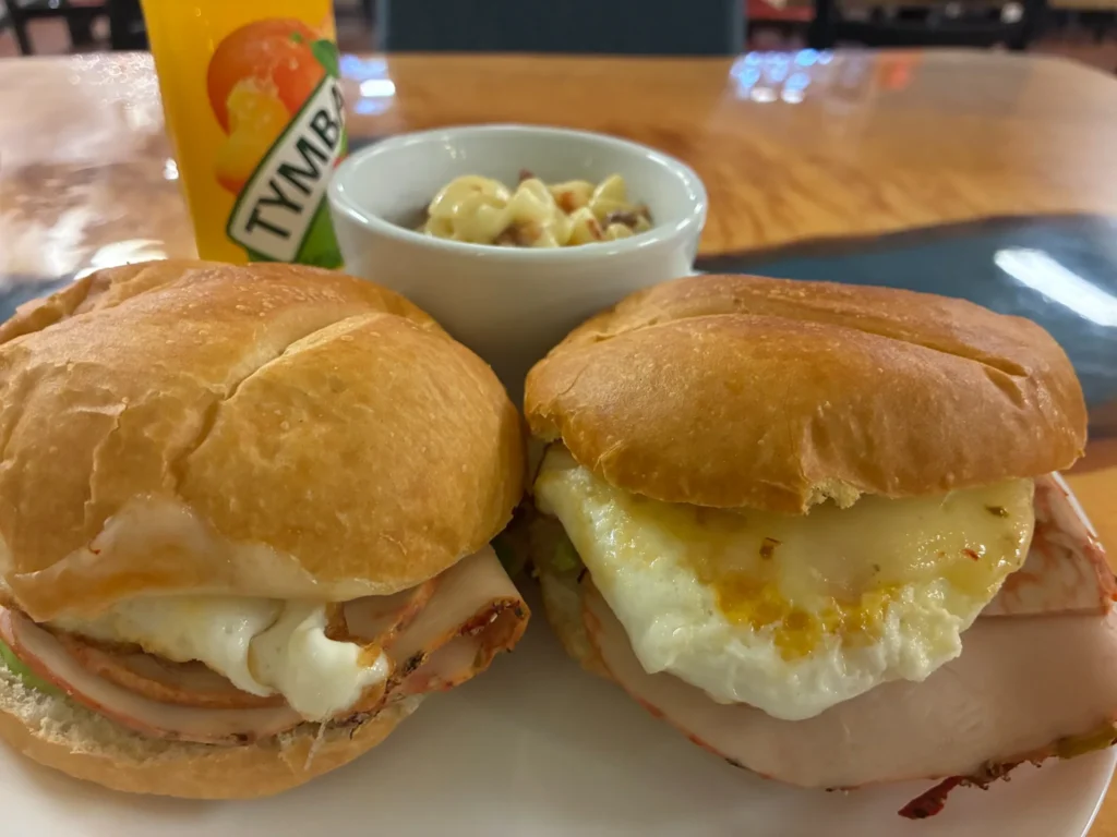 Two breakfast sandwiches with fried eggs and freshly sliced meats on fresh rolls served with a side of macaroni and cheese and fruit juice.