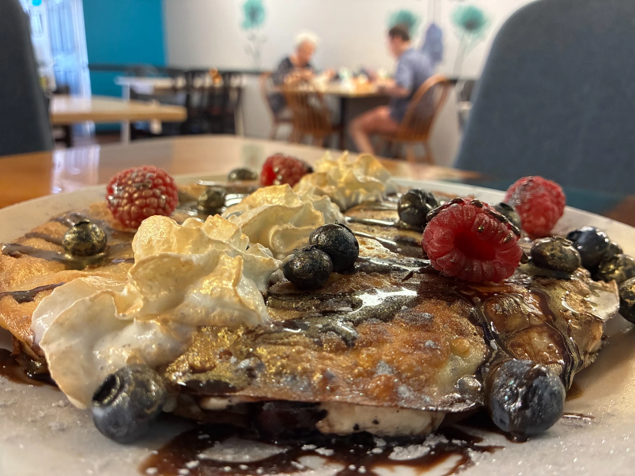 Double sweet cheese crepes served on a white platter, finished with chocolate drizzle, whipped cream, and fresh blackberries with customer dining in the background at one of the tables at the cafe-restaurant.