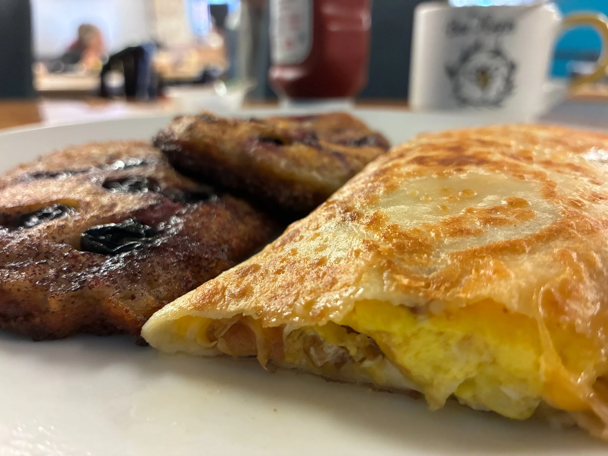 Breakfast plate with a ham-egg-and-cheese crepe in the foreground and two browned berry blinis alongside it with a cup of tea and ketchup behind it, and blurred customer dining at a table in the background.