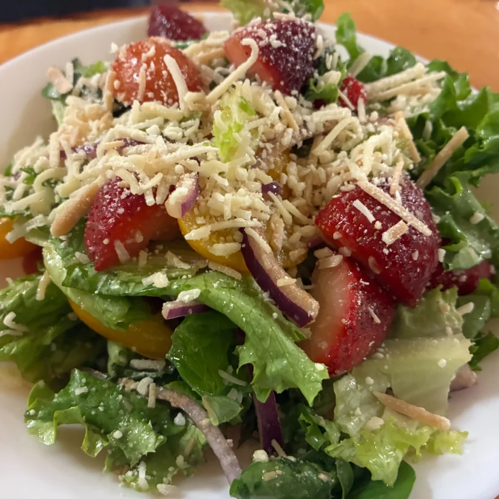 Garden salad with mixed greens, strawberries, cherry tomatoes, red onion, and grated cheese in a white bowl.