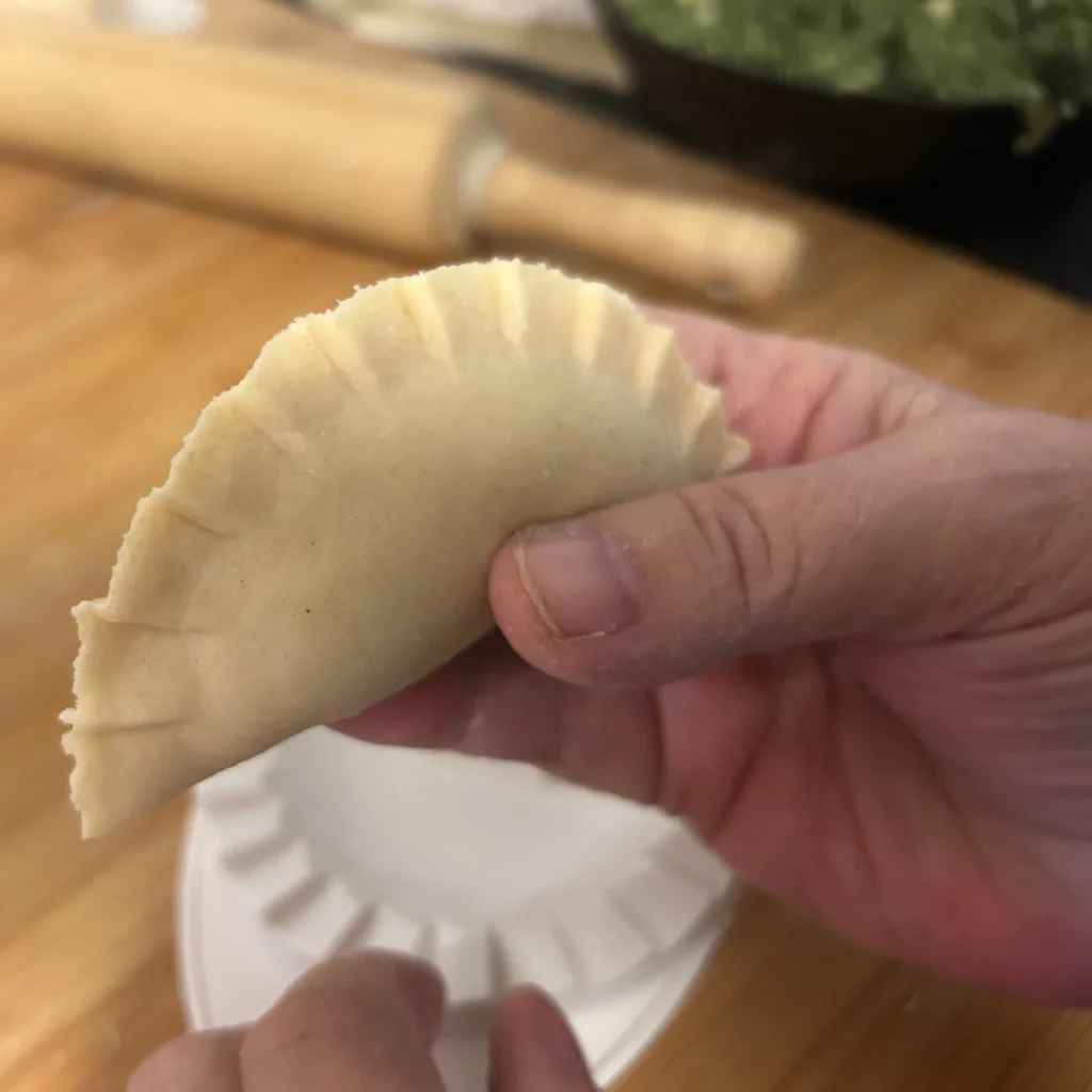 Hand holding a freshly crimped pierogi over a wooden prep board with a pierogi press and rolling pin in the background.