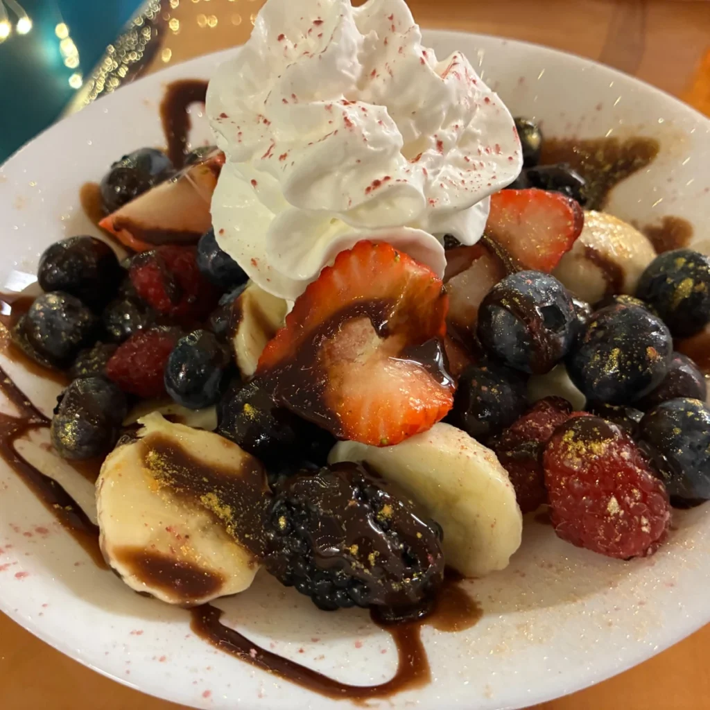 Fruit salad with banana, berries, chocolate drizzle, and whipped cream on a white plate.