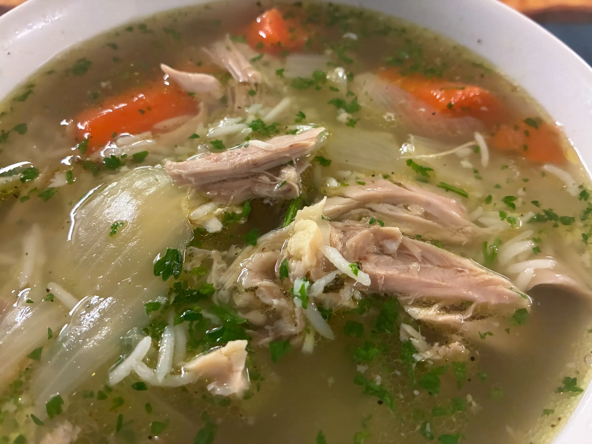 Clear chicken soup with shredded chicken, carrots, onion, herbs, and rice in a white bowl.
