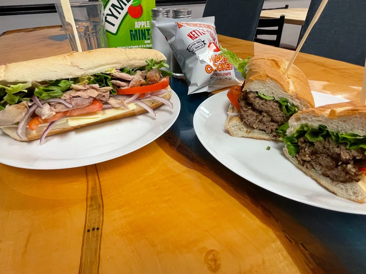 Grilled chicken sub and city chicken sub on white plates with chips at Kat's Café & Restaurant, with the city chicken sub cut in half and the grilled chicken sub topped with lettuce, tomato, red onion, and cheese. both served on white plates on the custom table in the dining room.