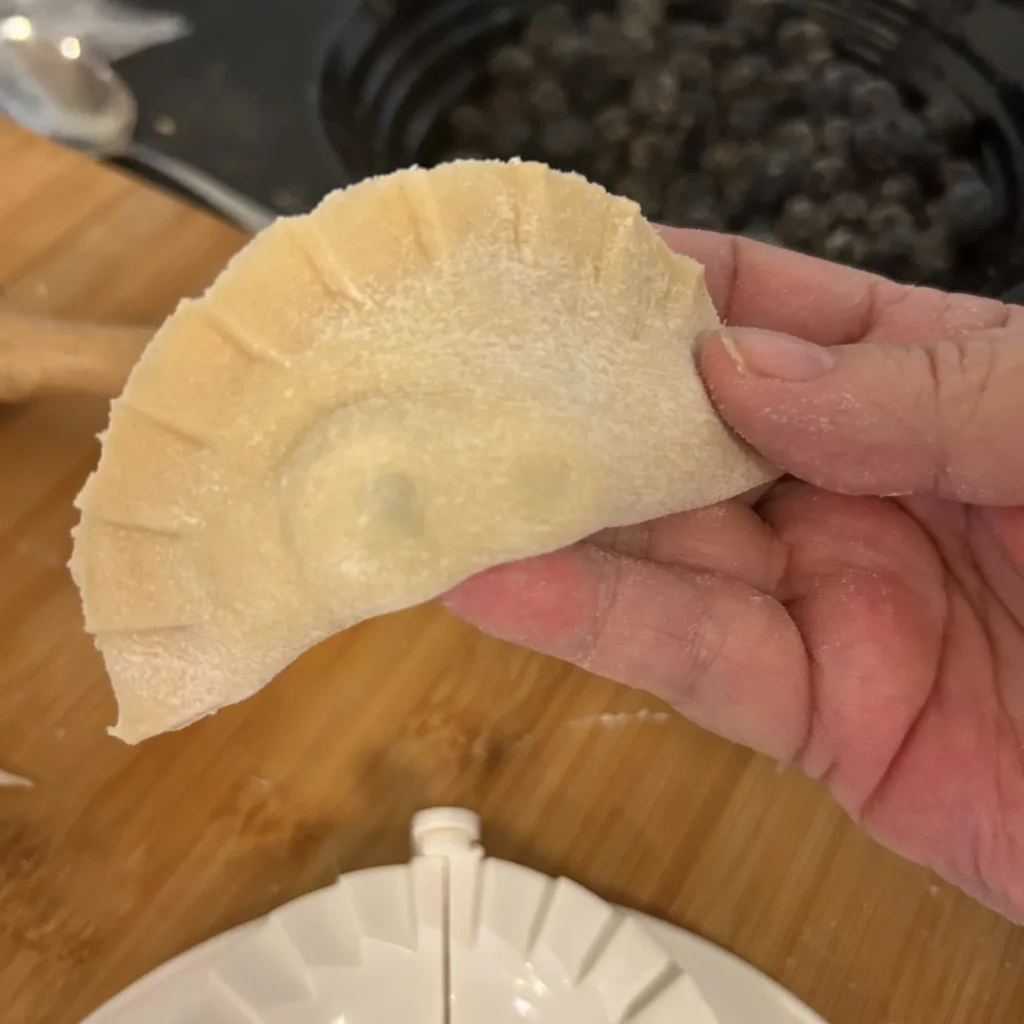 Hand holding a flour-dusted, hand-crimped pierogi made from scratch on a kitchen prep surface.