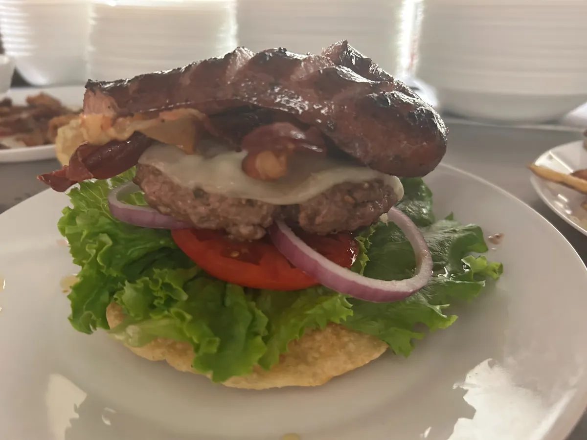 Pierogi smash burger layered with lettuce, tomato, red onion, melted cheese, bacon and kielbasa sausage on a plate in a kitchen setting with stacked plates in the background.