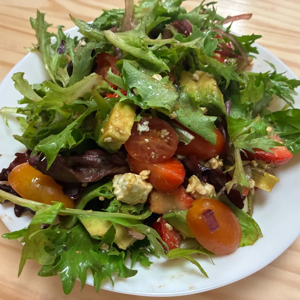 Leafy garden salad with avocado, cherry tomatoes, strawberries, and cheese on a white plate.