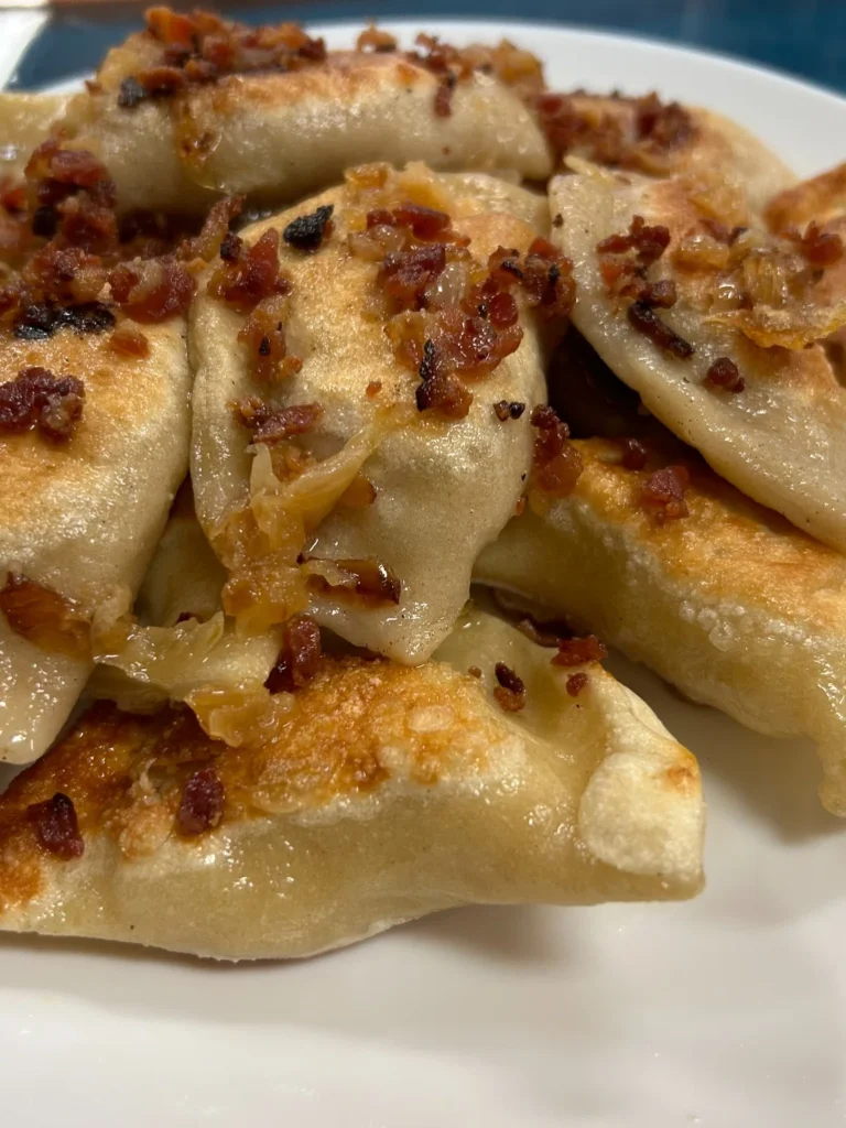 Pan-fried meat pierogies topped with sautéed onions and crumbled bacon on a white plate.
