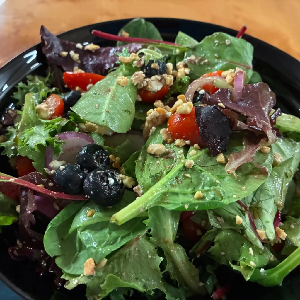 Mixed greens salad with blueberries, cherry tomatoes, red onion, and crunchy nuts in a black bowl.