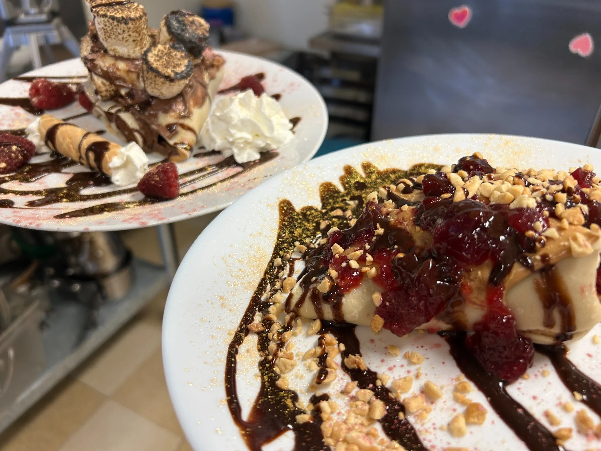 Foreground PB&J-style crepe topped with fruit jam, chopped nuts, and chocolate drizzle, with a toasted marshmallow dessert crepe behind it.