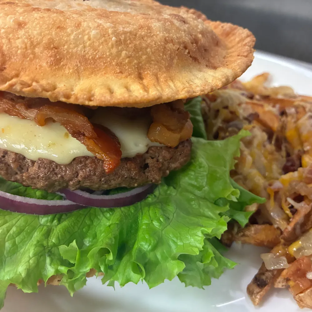 Pierogi burger with a crispy pierogi bun, bacon and melted cheese, served with loaded fries on a white plate.