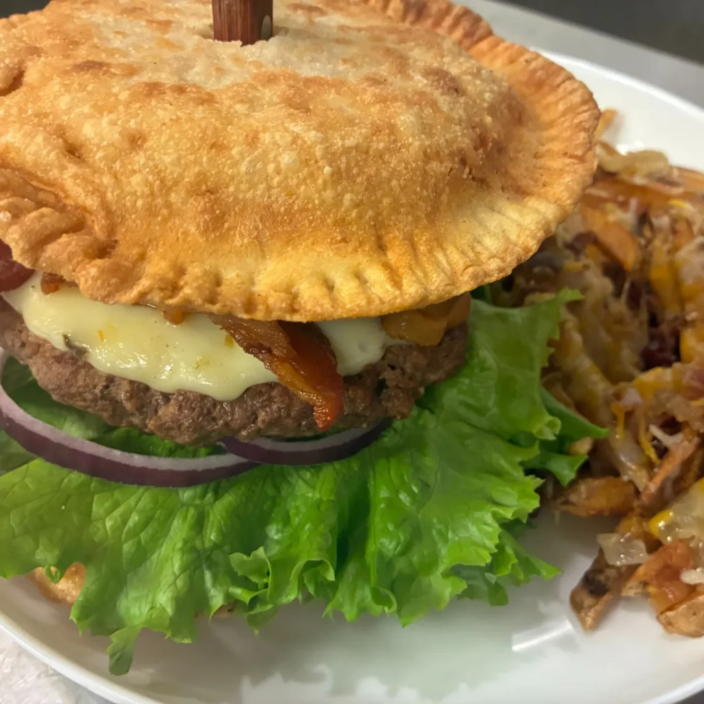 Pierogi smash burger with lettuce, onion, bacon, and melted cheese served with loaded fries on a white plate.